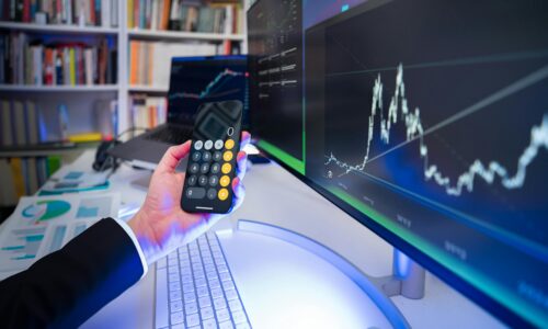 A person holding a mobile and there is a computer in front of him with him indoors at night, surrounded by technology, trading graphs, and music.