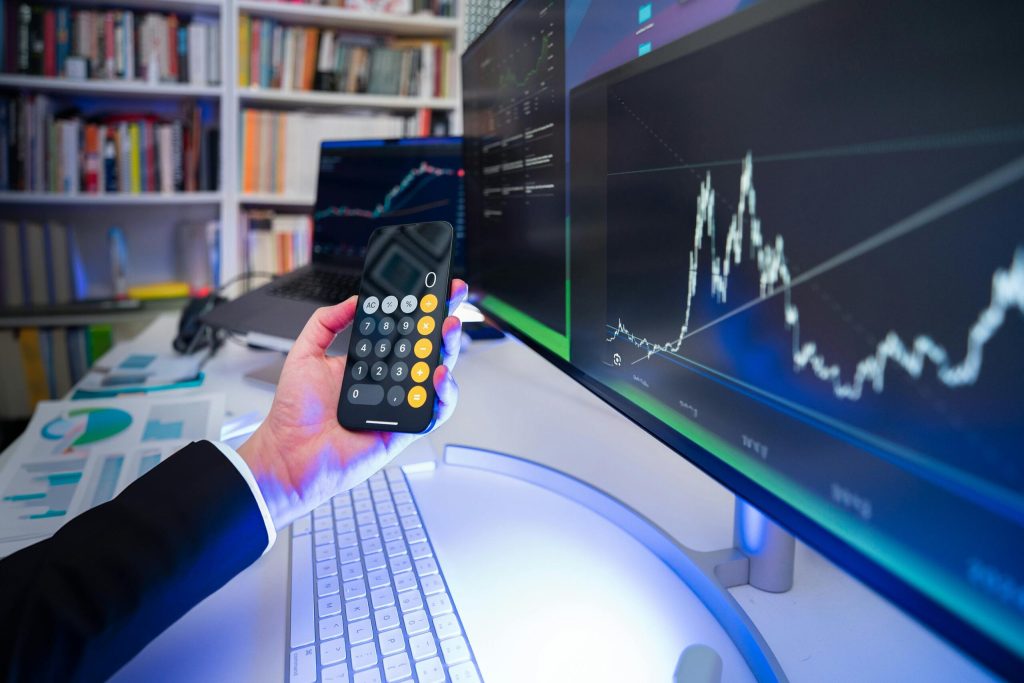 A person holding a mobile and there is a computer in front of him with him indoors at night, surrounded by technology, trading graphs, and music.
