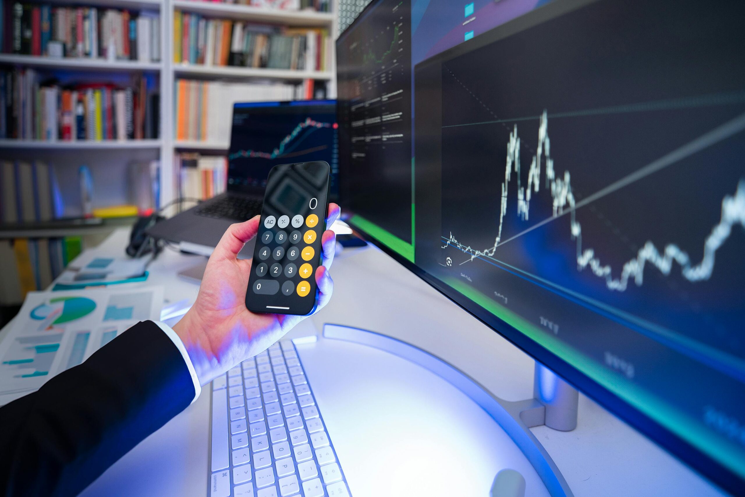 A person holding a mobile and there is a computer in front of him with him indoors at night, surrounded by technology, trading graphs, and music.